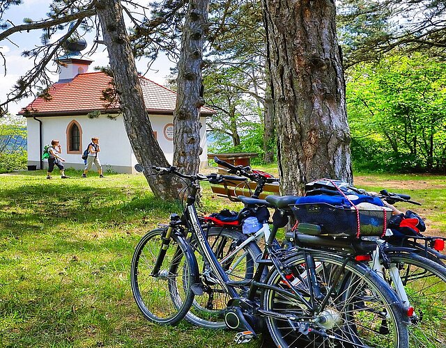 Aktivurlauber im Wald. Im Hintergrund eine Kapelle, davor Wanderer, im Vordergrund an einen Baum angelehnte Fahrräder.