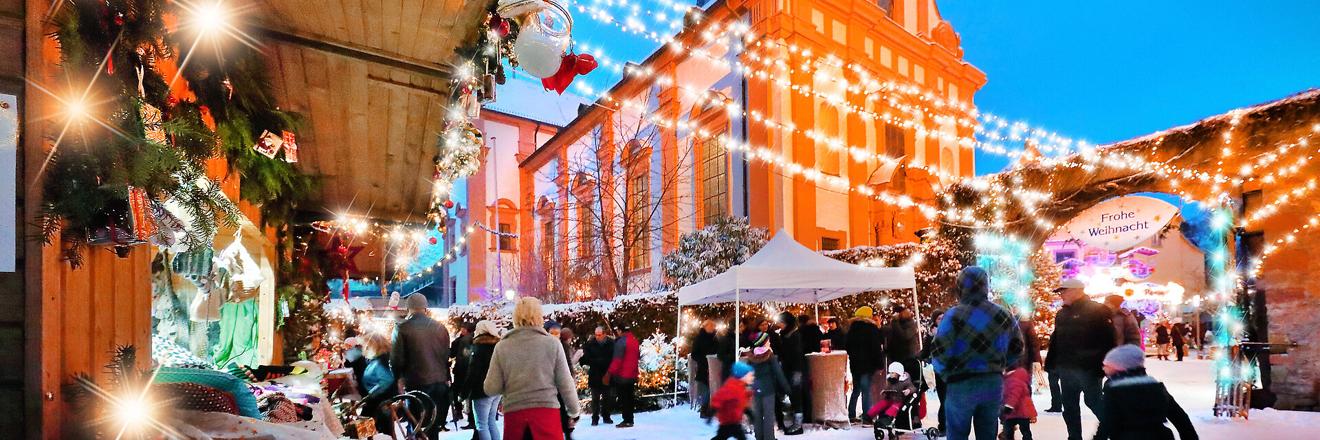 Veitshöchheimer Weihnachtsmarkt, im Vordergrund Buden und Besucher, im Hintergrund die illuminierte Kirche